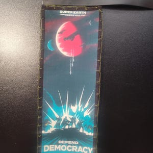 K&ouml;nnte beinhalten: Ein rechteckiger Aufn&auml;her mit dunkelblauem Rand und gen&auml;hter Kante. Der Aufn&auml;her zeigt ein Weltraum-Design mit einem roten Planeten, Raumschiffen und dem Text "SUPER EARTH NEEDS YOU!" und "DEFEND DEMOCRACY". Der Aufn&auml;her ist etwa 7,6 cm lang.