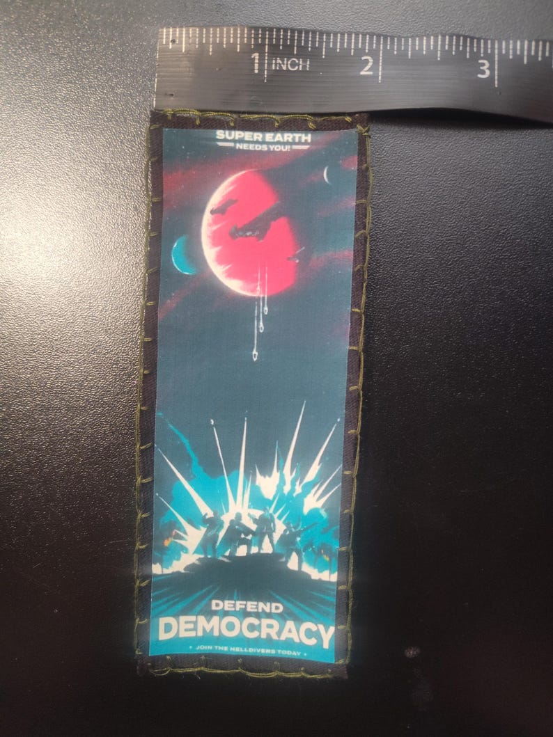 K&ouml;nnte beinhalten: Ein rechteckiger Aufn&auml;her mit dunkelblauem Rand und gen&auml;hter Kante. Der Aufn&auml;her zeigt ein Weltraum-Design mit einem roten Planeten, Raumschiffen und dem Text "SUPER EARTH NEEDS YOU!" und "DEFEND DEMOCRACY". Der Aufn&auml;her ist etwa 7,6 cm lang.