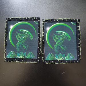 May include: Two square Alien-themed coasters. Each coaster features a green Xenomorph design against a dark background, framed by a black border with green stitching. The design includes a green halo and alien eggs.