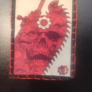 May include: A rectangular patch with a red skull design, a saw blade edge, and a gear emblem. The patch is edged with black fabric and orange stitching. The image has a gaming theme.