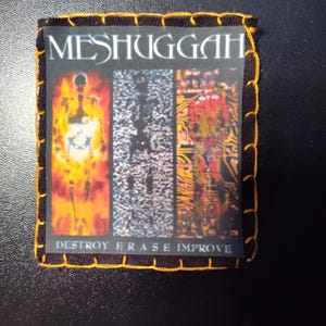 May include: A black patch with the band name "MESHUGGAH" and the words "DESTROY ERASE IMPROVE". The patch features a graphic with three vertical panels, each with a different image of a skeleton.