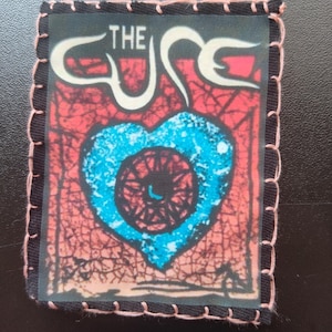 May include: A rectangular patch featuring the band name "THE CURE" in white script above a blue heart with a black spiral design. The background is red with a crackle effect. The patch has a black border with pink stitching.