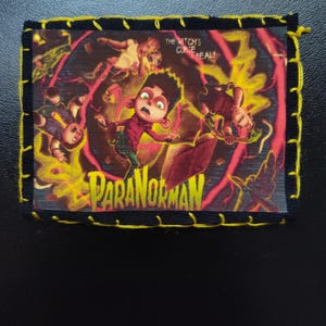 May include: A rectangular patch featuring the animated film "ParaNorman." The image shows a frightened boy surrounded by ghostly figures and yellow energy. The text "THE WITCH'S CURSE REAL!" and "PARANORMAN" are visible. The patch has a black border with yellow stitching.