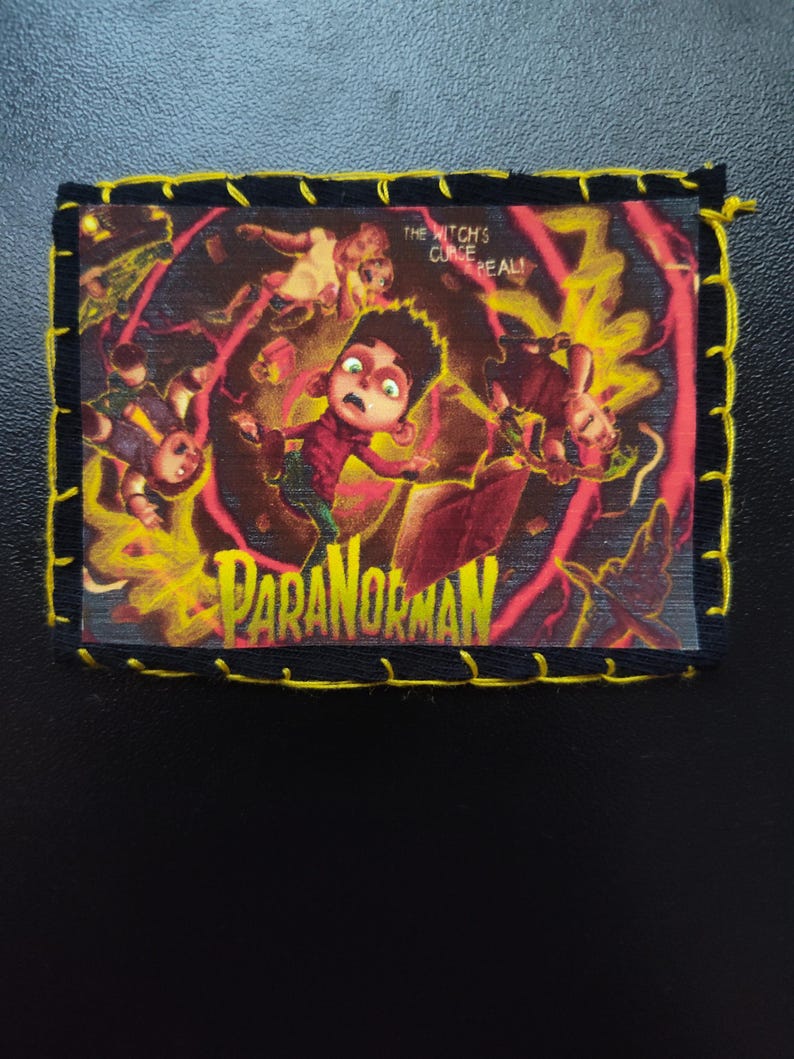 May include: A rectangular patch featuring the animated film "ParaNorman." The image shows a frightened boy surrounded by ghostly figures and yellow energy. The text "THE WITCH'S CURSE REAL!" and "PARANORMAN" are visible. The patch has a black border with yellow stitching.