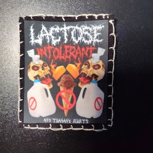 May include: Square patch with a black border and white stitching. The patch features the words "LACTOSE INTOLERANT" in a gothic font above two skull heads with milk jugs and cheese. A chocolate ice cream cone is crossed out. The text "MY TUMMY HURTS" is at the bottom.