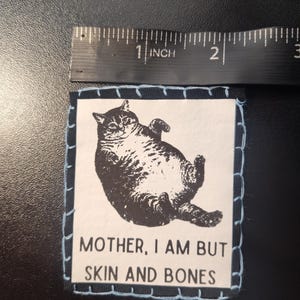 May include: A square patch with a black and white illustration of a cat lying on its back. The patch is approximately 2 inches wide and has a light blue stitched border. The text "MOTHER, I AM BUT SKIN AND BONES" is below the cat image.
