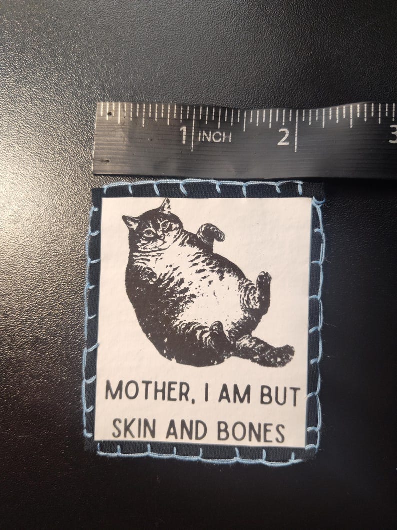 May include: A square patch with a black and white illustration of a cat lying on its back. The patch is approximately 2 inches wide and has a light blue stitched border. The text "MOTHER, I AM BUT SKIN AND BONES" is below the cat image.