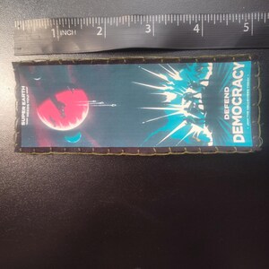 K&ouml;nnte beinhalten: Rechteckiger Aufn&auml;her mit Weltraum-Design. Der Aufn&auml;her zeigt einen roten Planeten, Raumschiffe und den Text "SUPER EARTH NEEDS YOU!" und "DEFEND DEMOCRACY". Der Aufn&auml;her ist etwa 12,7 cm lang.