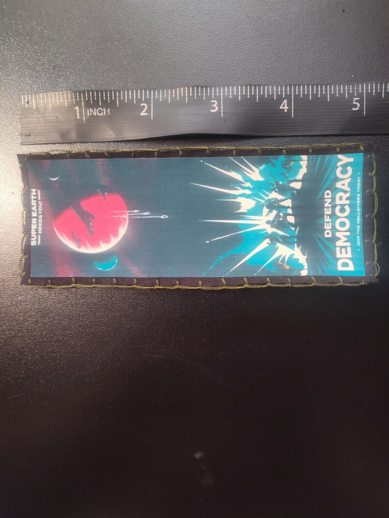 K&ouml;nnte beinhalten: Rechteckiger Aufn&auml;her mit Weltraum-Design. Der Aufn&auml;her zeigt einen roten Planeten, Raumschiffe und den Text "SUPER EARTH NEEDS YOU!" und "DEFEND DEMOCRACY". Der Aufn&auml;her ist etwa 12,7 cm lang.