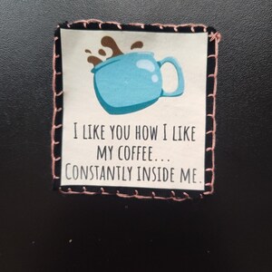 K&ouml;nnte beinhalten: Ein Stoffaufn&auml;her mit einem hellblauen Kaffeetassen-Design und dem Text "I LIKE YOU HOW I LIKE MY COFFEE... CONSTANTLY INSIDE ME." Der Aufn&auml;her hat einen schwarzen Rand mit rosa N&auml;hten.