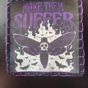 Peut inclure: Écusson carré avec une bordure noire et des coutures violettes. L'écusson représente un papillon de nuit avec une tête de mort, des flammes et des chauves-souris, avec les mots "MAKE THEM SUFFER" en violet.