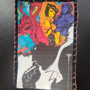 May include: A rectangular patch featuring a colorful anime-style illustration. The design includes figures in shades of red, yellow, and blue, with a black and white silhouette of a person smoking a cigarette. The patch is bordered with brown stitching.