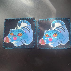 May include: Two square patches with a blue tiger design. The tiger has orange eyes, a pink tongue, and a white and blue striped tail. The patches are black with blue stitching around the edges.