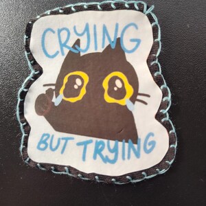May include: A fabric patch featuring a cartoon black cat with yellow eyes and tears, with the words "CRYING BUT TRYING" in blue. The patch has a white background and a black border with blue stitching.