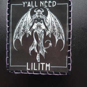 May include: Black square patch featuring a white illustration of a winged figure, with the text "Y'ALL NEED" above and "LILITH" below. The patch has a black border with purple stitching.