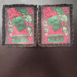 May include: Two square patches with the text "THE EVIL DEAD" in pink and green. The patches feature a movie poster design with green and red imagery and are framed with black fabric and green stitching.