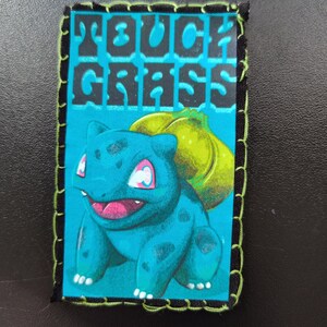 May include: A rectangular patch with a teal background featuring a cartoon Bulbasaur and the words "TOUCH GRASS" in black. The patch is bordered with black fabric and green stitching.