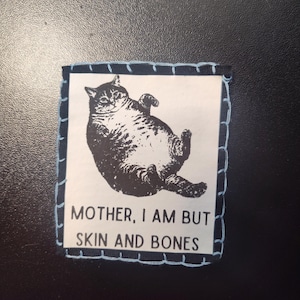 May include: A square patch with a black and white illustration of a cat lying on its back. The patch has a light blue stitched border and the text "MOTHER, I AM BUT SKIN AND BONES".