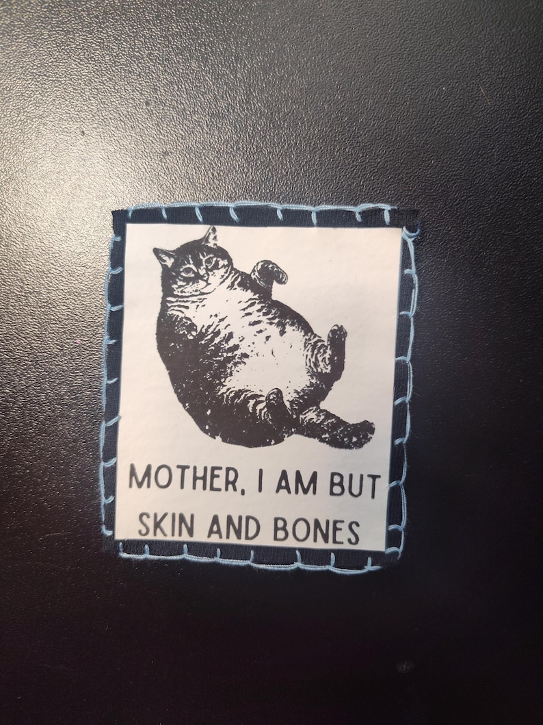 May include: A square patch with a black and white illustration of a cat lying on its back. The patch has a light blue stitched border and the text "MOTHER, I AM BUT SKIN AND BONES".