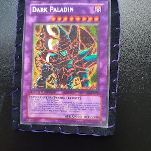 May include: A Dark Paladin trading card with a purple background, encased in a black fabric border with purple stitching. The card features a detailed illustration of a warrior and text that reads "DARK PALADIN".