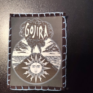 May include: A rectangular patch featuring the word "GOJIRA" above a circular design of a volcano erupting with a sun face. The patch has a black border with light blue stitching.