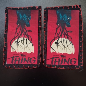 Peut inclure: Deux écussons rectangulaires avec un fond rouge, représentant un monstre et le texte "THE THING". Les bordures sont noires avec des coutures rouges.