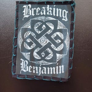 May include: A rectangular patch with the words "Breaking Benjamin" in a gothic font. The patch features a white and black Celtic knot design. The edges are stitched with teal thread.