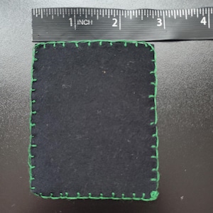 K&ouml;nnte beinhalten: Ein quadratischer, schwarzer Stoffartikel mit gr&uuml;nen N&auml;hten an den R&auml;ndern. Der Artikel misst etwa 7,6 cm im Durchmesser. Die N&auml;hte sind hellgr&uuml;n und stehen im Kontrast zum dunklen Stoff.