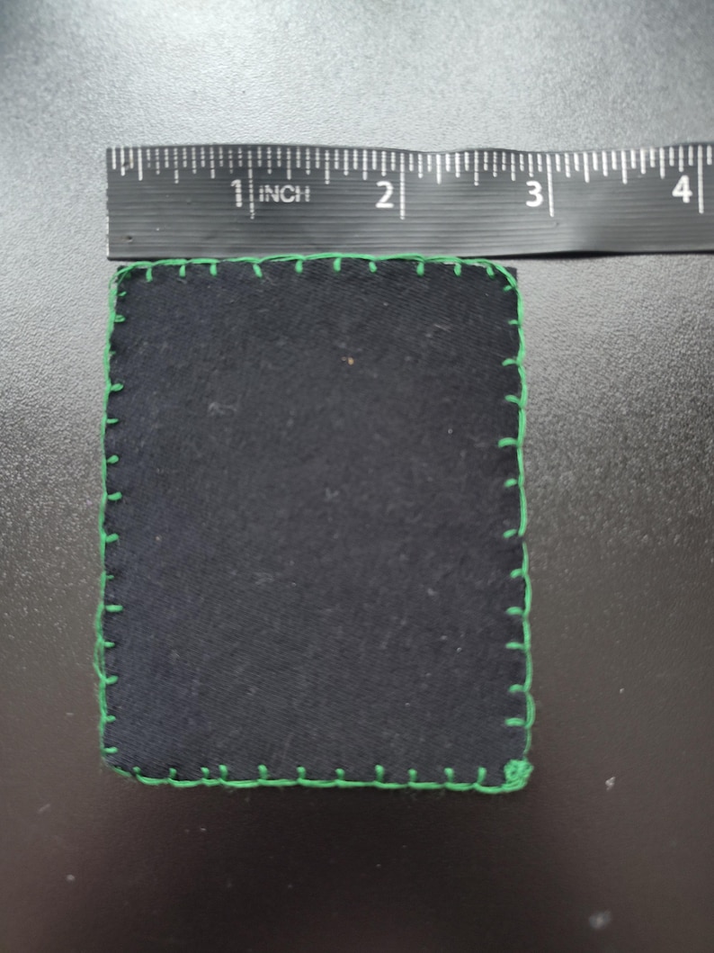 K&ouml;nnte beinhalten: Ein quadratischer, schwarzer Stoffartikel mit gr&uuml;nen N&auml;hten an den R&auml;ndern. Der Artikel misst etwa 7,6 cm im Durchmesser. Die N&auml;hte sind hellgr&uuml;n und stehen im Kontrast zum dunklen Stoff.