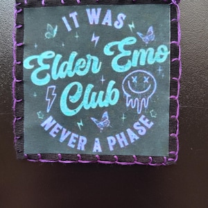 May include: A square, black patch with purple stitching around the edges. The patch features the text "IT WAS Elder Emo Club NEVER A PHASE" in teal and purple, along with butterfly and lightning bolt graphics.