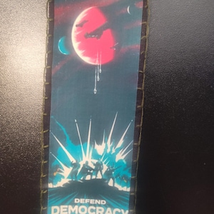 May include: Rectangular patch with a space-themed design. The top reads "SUPER EARTH NEEDS YOU!" above a red planet with spacecraft. The bottom says "DEFEND DEMOCRACY" with a blue explosion graphic and silhouetted figures.