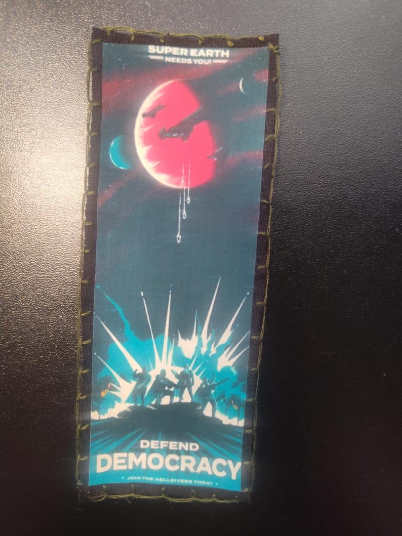 K&ouml;nnte beinhalten: Rechteckiger Aufn&auml;her mit Weltraum-Design. Oben steht "SUPER EARTH NEEDS YOU!" &uuml;ber einem roten Planeten mit Raumschiffen. Unten steht "DEFEND DEMOCRACY" mit einer blauen Explosionsgrafik und Silhouetten.