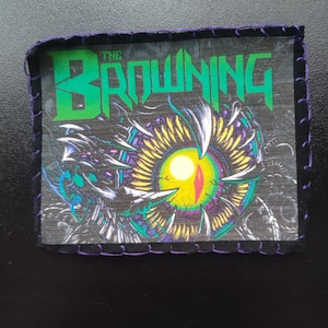 May include: A rectangular patch with the text "THE BROWNING" in green block letters. The patch features a detailed illustration of a stylized eye with yellow, purple, and white elements, and is outlined with purple stitching.
