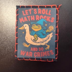 May include: A square patch with a black border and red stitching. The patch features a cartoon frog in a wizard hat riding a duck, with the text "LET'S ROLL MATH ROCKS AND DO WAR CRIMES" in red and white.