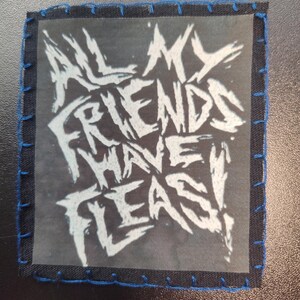 May include: Square patch with the text "ALL MY FRIENDS HAVE FLEAS!" in white, stylized lettering. The text is on a transparent background, surrounded by a black fabric border with blue stitching.