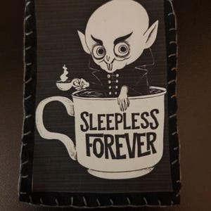 May include: A black and white patch featuring a cartoon vampire in a coffee cup. The vampire is holding a small cup of coffee. The text on the cup reads "SLEEPLESS FOREVER". The patch has a black border with white stitching.