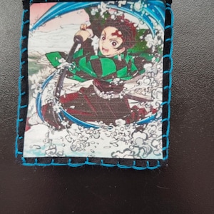 May include: A rectangular patch featuring an anime character in a dynamic pose with a sword, surrounded by water effects. The image is framed with black fabric and stitched with blue thread.