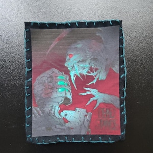 May include: A rectangular patch with a monster and a figure in a space suit. The artwork is in blue, red, and grey. The patch has a black border with teal stitching and the text "DEAD SPACE".