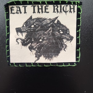 May include: A square patch with the text "EAT THE RICH" above a black and white illustration of three snarling wolf-like creatures. The patch has a black border with green stitching.