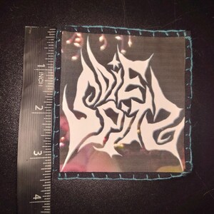 K&ouml;nnte beinhalten: Quadratischer Aufn&auml;her mit wei&szlig;em, abstraktem Design auf schwarzem Hintergrund, misst ca. 5 cm x 5 cm. Der Aufn&auml;her hat einen t&uuml;rkisfarbenen Rand mit t&uuml;rkisfarbenen N&auml;hten. Das Design scheint ein stilisiertes Logo zu sein.