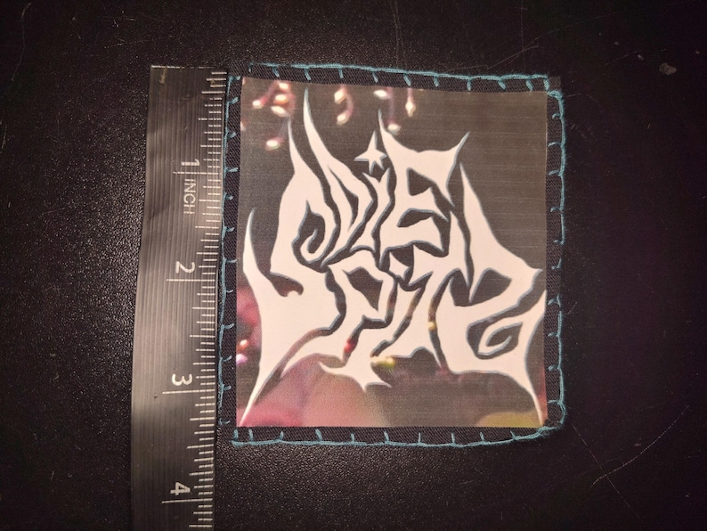 K&ouml;nnte beinhalten: Quadratischer Aufn&auml;her mit wei&szlig;em, abstraktem Design auf schwarzem Hintergrund, misst ca. 5 cm x 5 cm. Der Aufn&auml;her hat einen t&uuml;rkisfarbenen Rand mit t&uuml;rkisfarbenen N&auml;hten. Das Design scheint ein stilisiertes Logo zu sein.