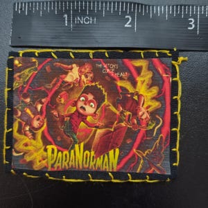 May include: A rectangular patch featuring the movie title "ParaNorman" with a distressed design. The patch has a black border with yellow stitching. The image includes the text "The Witch's Curse is Real!" and a ruler showing the patch is approximately 2.5 inches wide.