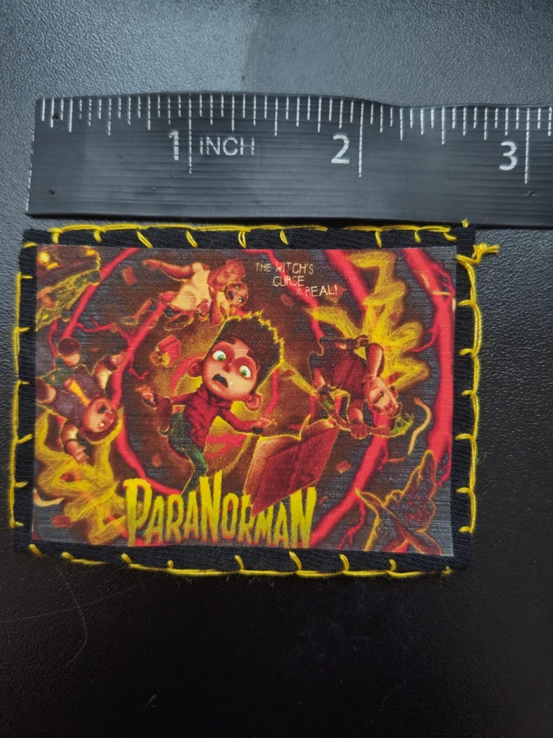 May include: A rectangular patch featuring the movie title "ParaNorman" with a distressed design. The patch has a black border with yellow stitching. The image includes the text "The Witch's Curse is Real!" and a ruler showing the patch is approximately 2.5 inches wide.