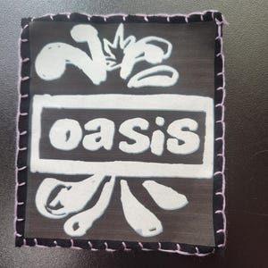 May include: Square black patch with white text and design. The word "oasis" is in a white rectangle. The patch has a white design above the word "oasis" and below it. The patch is outlined with pink stitching.