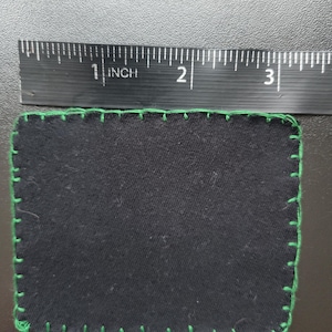 K&ouml;nnte beinhalten: Ein rechteckiger schwarzer Stoffflicken mit gr&uuml;nen N&auml;hten an den R&auml;ndern. Der Flicken ist etwa 7,6 cm lang und 5,1 cm breit. Die gr&uuml;nen N&auml;hte bilden eine dekorative Umrandung.