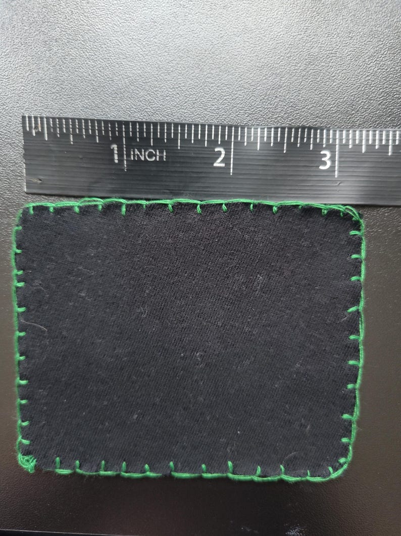 K&ouml;nnte beinhalten: Ein rechteckiger schwarzer Stoffflicken mit gr&uuml;nen N&auml;hten an den R&auml;ndern. Der Flicken ist etwa 7,6 cm lang und 5,1 cm breit. Die gr&uuml;nen N&auml;hte bilden eine dekorative Umrandung.