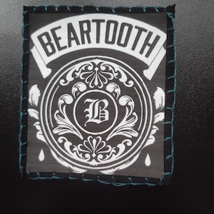 May include: Square patch with the word "BEARTOOTH" in white on a black and white circular design. The patch has a black border with teal stitching.