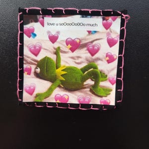 May include: A square patch featuring a green frog character lying down, surrounded by pink hearts. The text "love u soooooOoOo much" is printed at the top. The patch has a black border with pink stitching.
