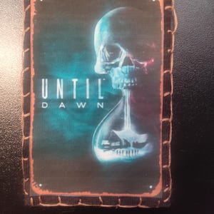 May include: Rectangular patch featuring the text "UNTIL DAWN" above an image of a skull with an hourglass. The skull is white and blue, with a dark background. The patch has a brown border with decorative stitching.