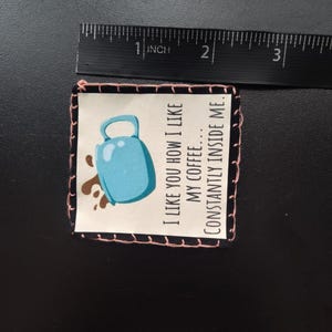 K&ouml;nnte beinhalten: Ein kleines, quadratisches Patch mit schwarzem Rand und rosa N&auml;hten. Das Patch zeigt eine Illustration einer hellblauen Kaffeetasse mit braunen Kaffeespritzern. Der Text lautet: "I LIKE YOU HOW I LIKE MY COFFEE... CONSTANTLY INSIDE ME."